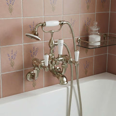 A vintage-style brushed nickel bath tap set with white lever handles and a matching handheld shower attachment, mounted on a pink floral tile wall above a white bathtub.
