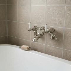 Brushed nickel tradiational bath filler with white ceramic handles mounted on a beige tiled wall above a white bathtub, with a small soap dish holding a bar of soap.